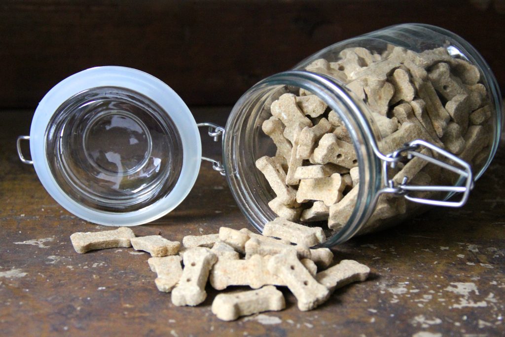 This is a glass mason jar with rubber seal set to the right with dog cookies pouring out onto a wooden shelf in the daylight.
